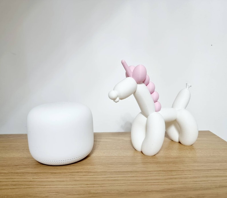 May include: A white ceramic unicorn figurine with pink balloon-like details. The unicorn is standing on a wooden surface next to a white round smart home device.