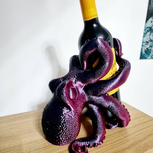 May include: A purple and black octopus shaped wine bottle holder. The octopus has eight tentacles and is holding a bottle of red wine.