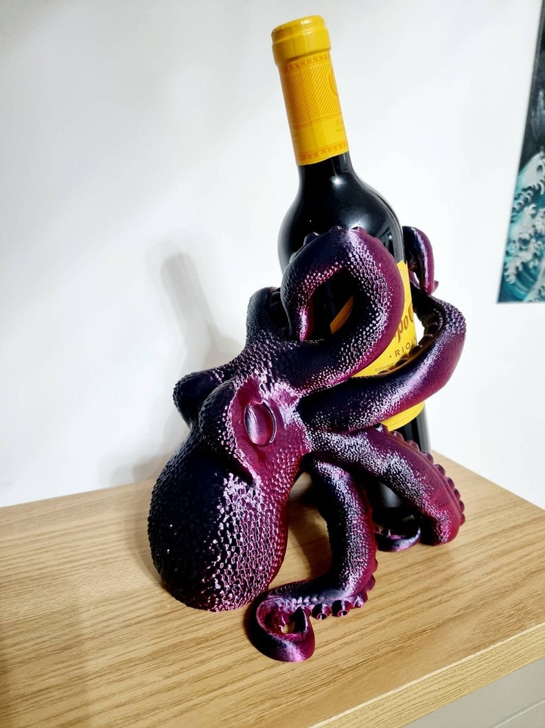 May include: A purple and black octopus shaped wine bottle holder. The octopus has eight tentacles and is holding a bottle of red wine.