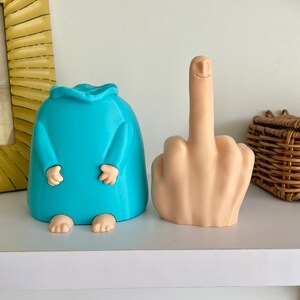May include: Two 3D printed figurines. One is a blue, humanoid figure with arms and legs. The other is a nude, fleshy hand with a smiling face giving the middle finger.