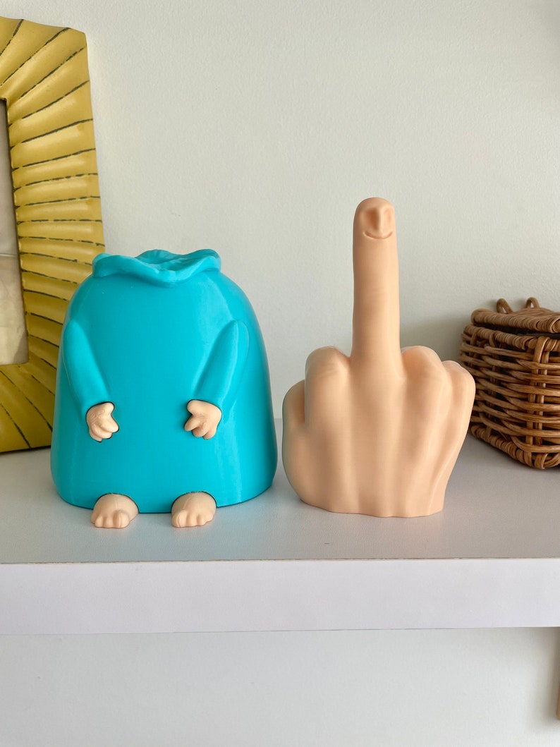 May include: Two 3D printed figurines. One is a blue, humanoid figure with arms and legs. The other is a nude, fleshy hand with a smiling face giving the middle finger.
