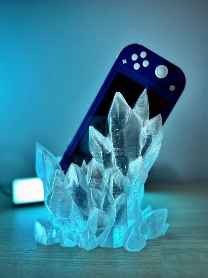 May include: A blue Nintendo Switch console is resting in a clear crystal-shaped stand. The stand is made of translucent plastic and resembles a cluster of ice crystals.