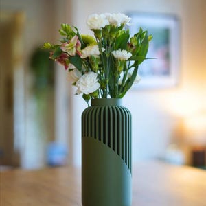 May include: A green vase with a unique design, holding a bouquet of white and green flowers. The vase has a cylindrical shape with vertical lines on one side and a smooth, angled panel on the other. The flowers include white carnations.