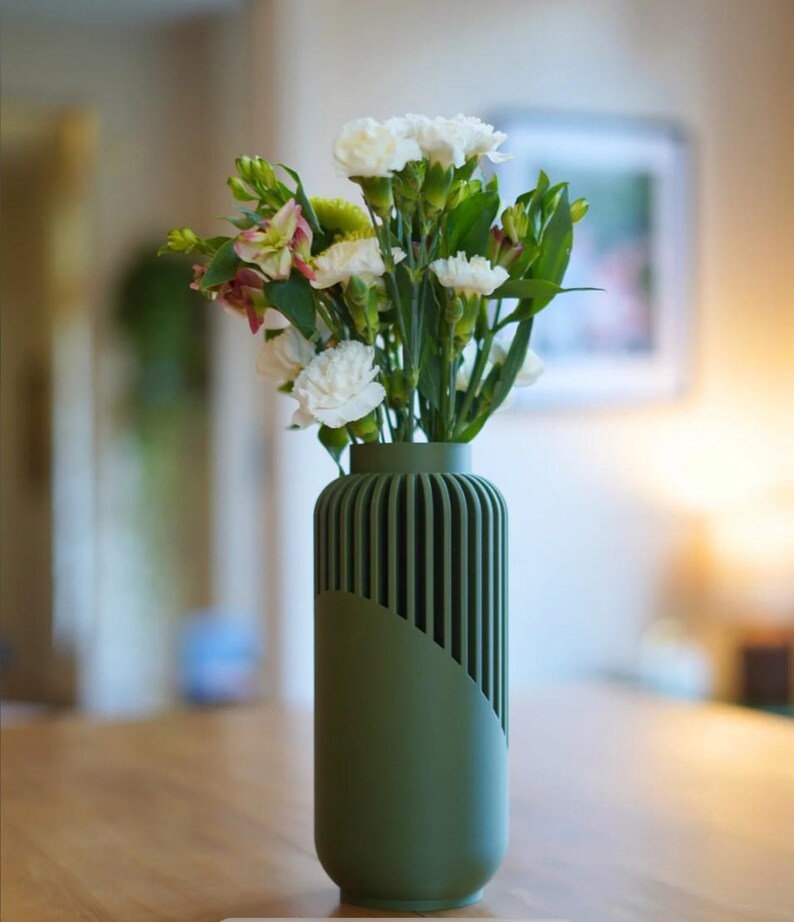May include: A green vase with a unique design, holding a bouquet of white and green flowers. The vase has a cylindrical shape with vertical lines on one side and a smooth, angled panel on the other. The flowers include white carnations.