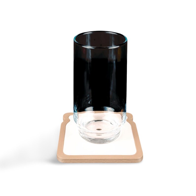 May include: A clear glass tumbler with a black interior sits atop a small, clear glass bowl. The bowl rests on a coaster shaped like a slice of bread, with a white center and a light brown border. The items are set against a white background.