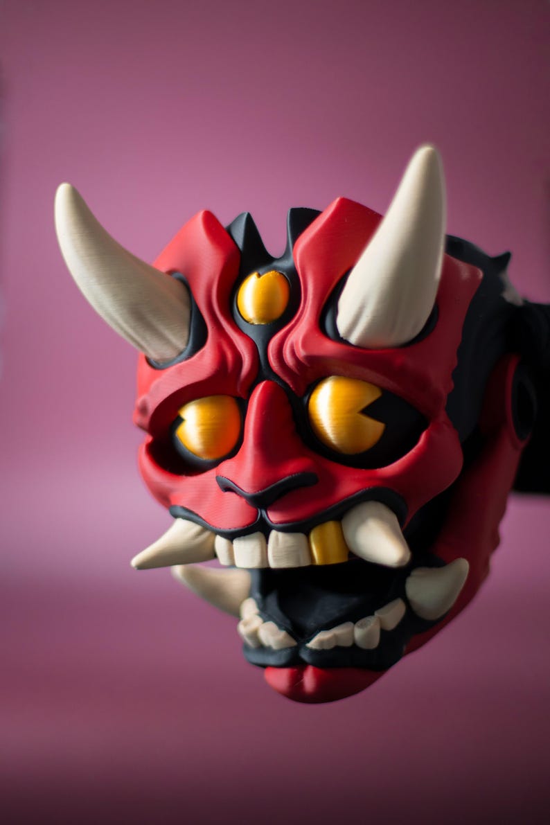 May include: A red and black Oni mask with three eyes and large white horns. The mask has a wide open mouth with white and gold teeth. The background is a solid pink color.