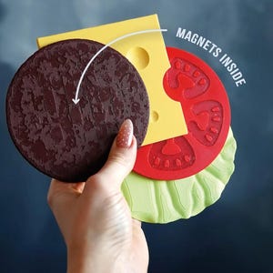 Op de afbeelding: Een gelaagde, burger-vormige magneetset. De set bevat een getextureerde bruine patty, gele kaas, rode tomaat en groene sla. De tekst "MAGNETS INSIDE" is zichtbaar.
