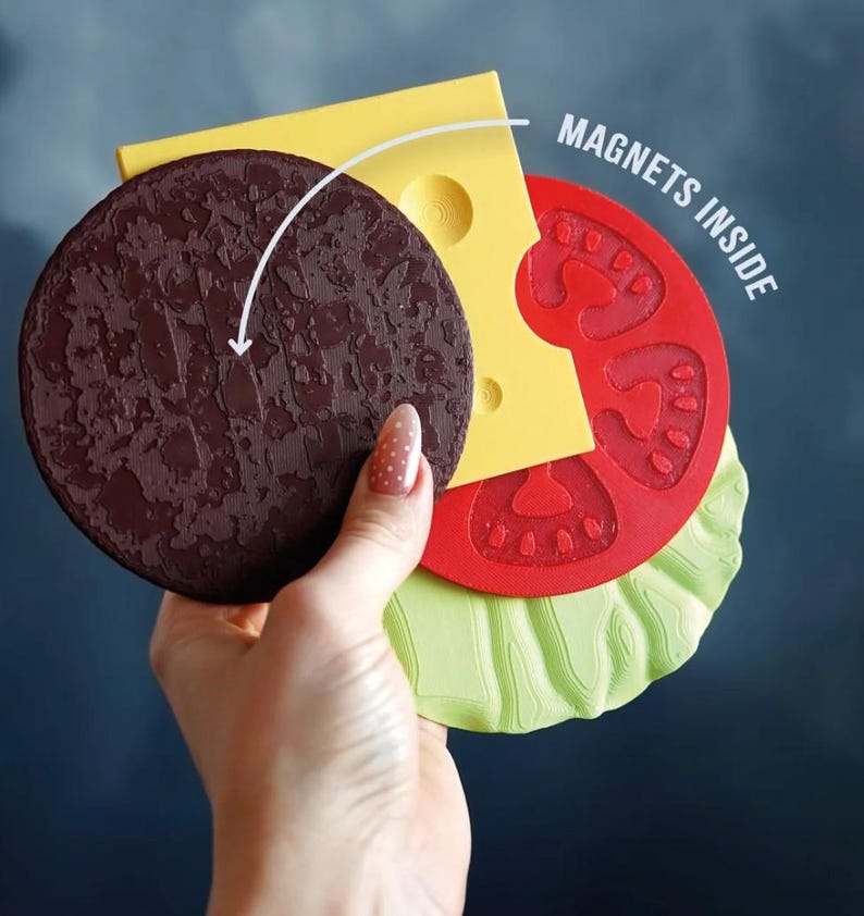Op de afbeelding: Een gelaagde, burger-vormige magneetset. De set bevat een getextureerde bruine patty, gele kaas, rode tomaat en groene sla. De tekst "MAGNETS INSIDE" is zichtbaar.