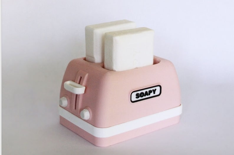 K&ouml;nnte beinhalten: Ein rosa Toaster mit zwei wei&szlig;en Seifenst&uuml;cken darin. Der Toaster hat einen wei&szlig;en Rand und das Wort "SOAPY" ist auf der Vorderseite aufgedruckt.