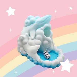 Puede incluir: Una bandeja de dados caprichosa en forma de nube, en blanco y azul claro. Dos dados con puntos negros descansan en una piscina azul en la base. El fondo presenta un arco&iacute;ris y estrellas.