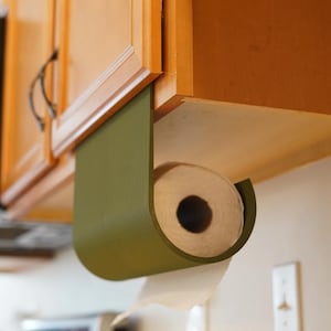 May include: An olive green paper towel holder is mounted under a wooden cabinet. The holder has a curved design, and a roll of white paper towels is visible. The holder is designed to dispense paper towels.