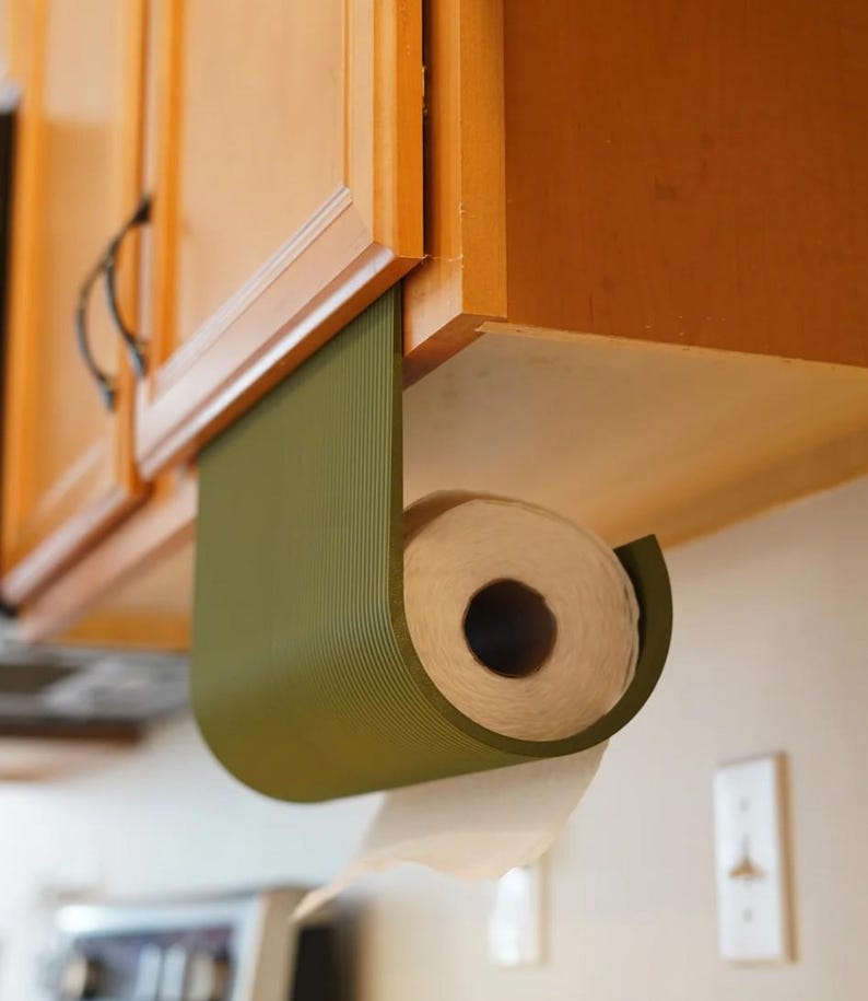 May include: An olive green paper towel holder is mounted under a wooden cabinet. The holder has a curved design, and a roll of white paper towels is visible. The holder is designed to dispense paper towels.