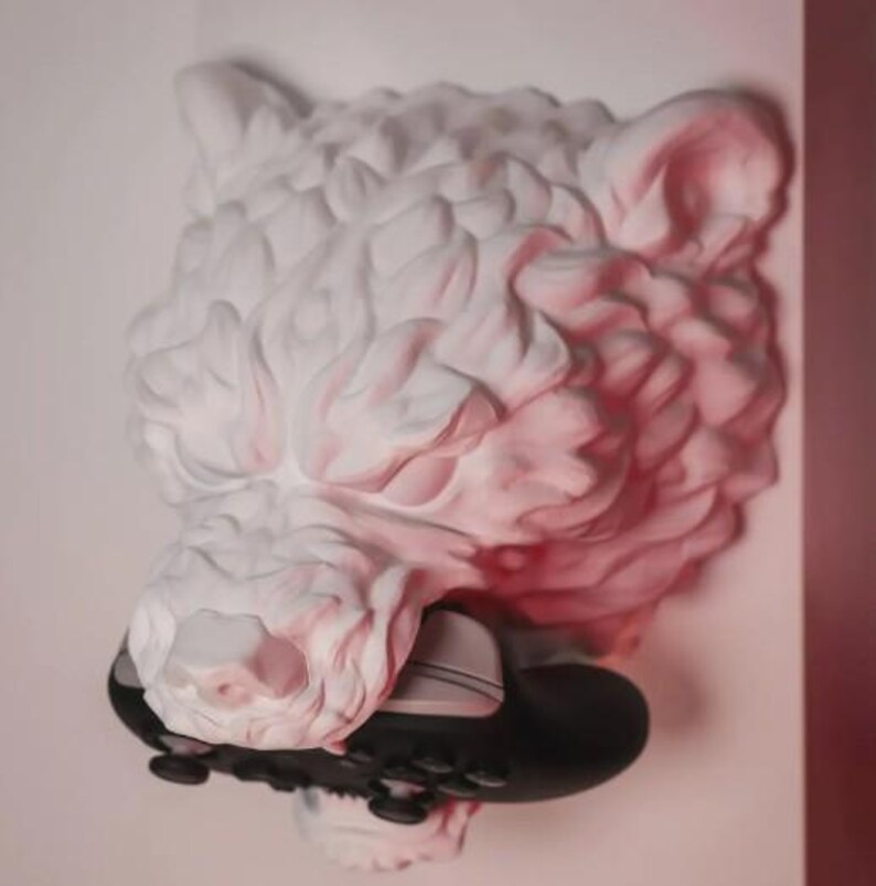 May include: A white, textured sculpture of a bear's head with a black and gray game controller in its mouth. The sculpture is mounted on a white wall, with a hint of pink lighting.
