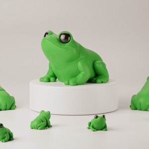 May include: A collection of green frog figurines. One large frog sits atop a white pedestal, surrounded by smaller frog figures. The frogs have large, black eyes and a glossy finish, creating a playful and whimsical aesthetic.