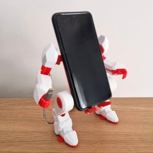 May include: A white and red robot phone stand with a black smartphone in the holder. The robot has a cable attached to it.
