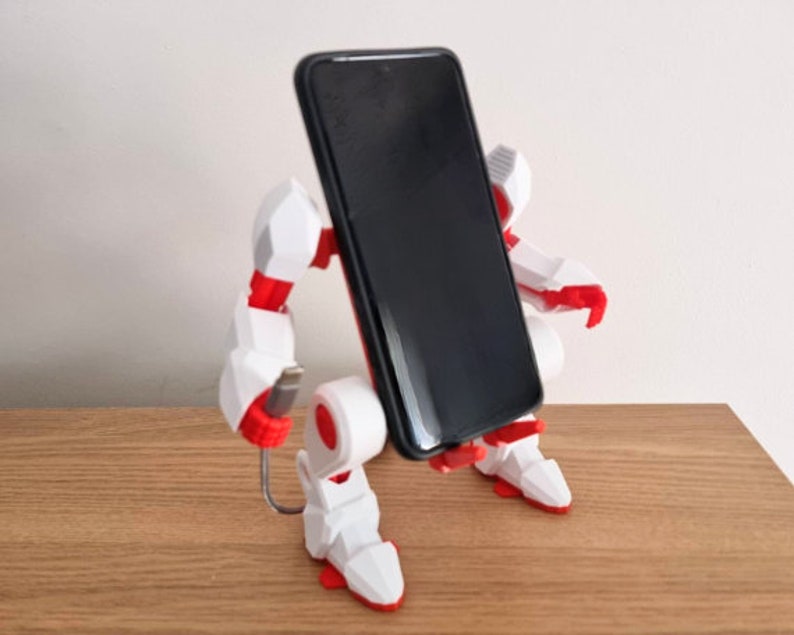 May include: A white and red robot phone stand with a black smartphone in the holder. The robot has a cable attached to it.
