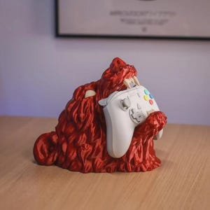 May include: A red, monster-shaped controller holder with a white controller. The holder has sharp teeth and a textured surface. The controller has colored buttons and a directional pad. The holder is on a wooden surface.