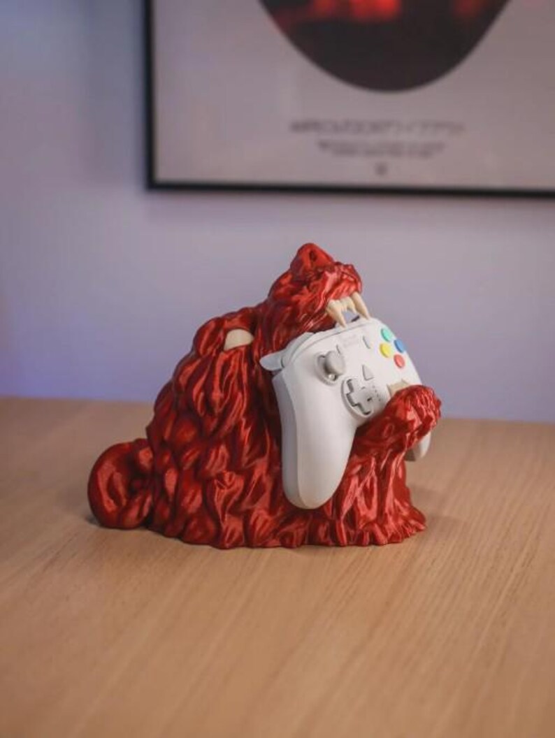 May include: A red, monster-shaped controller holder with a white controller. The holder has sharp teeth and a textured surface. The controller has colored buttons and a directional pad. The holder is on a wooden surface.