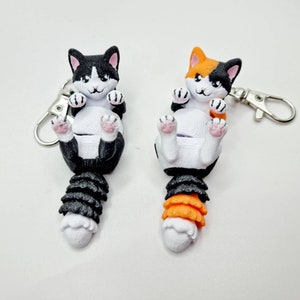 K&ouml;nnte beinhalten: Zwei Schl&uuml;sselanh&auml;nger in Form von Katzen, einer schwarz-wei&szlig;, der andere orange-wei&szlig;, mit baumelnden Schw&auml;nzen. Die Katzen sind in einer zusammengerollten Position mit ihren Pfoten in der Luft.