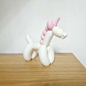 May include: A white balloon animal shaped like a unicorn with a pink mane and horn. The unicorn is standing on a wooden surface.