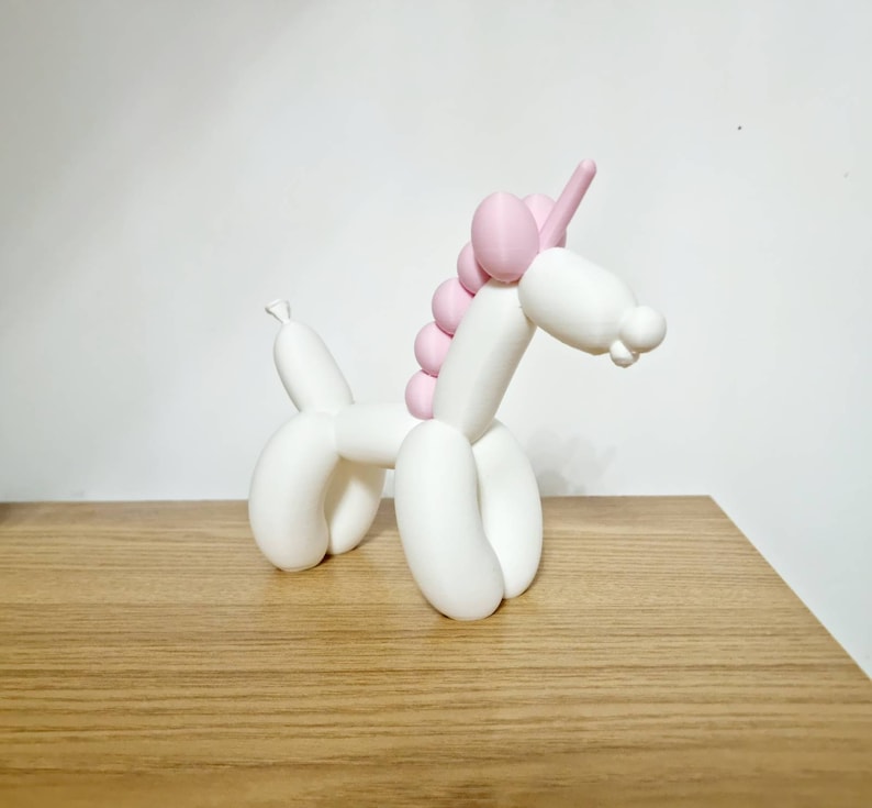 May include: A white balloon animal shaped like a unicorn with a pink mane and horn. The unicorn is standing on a wooden surface.