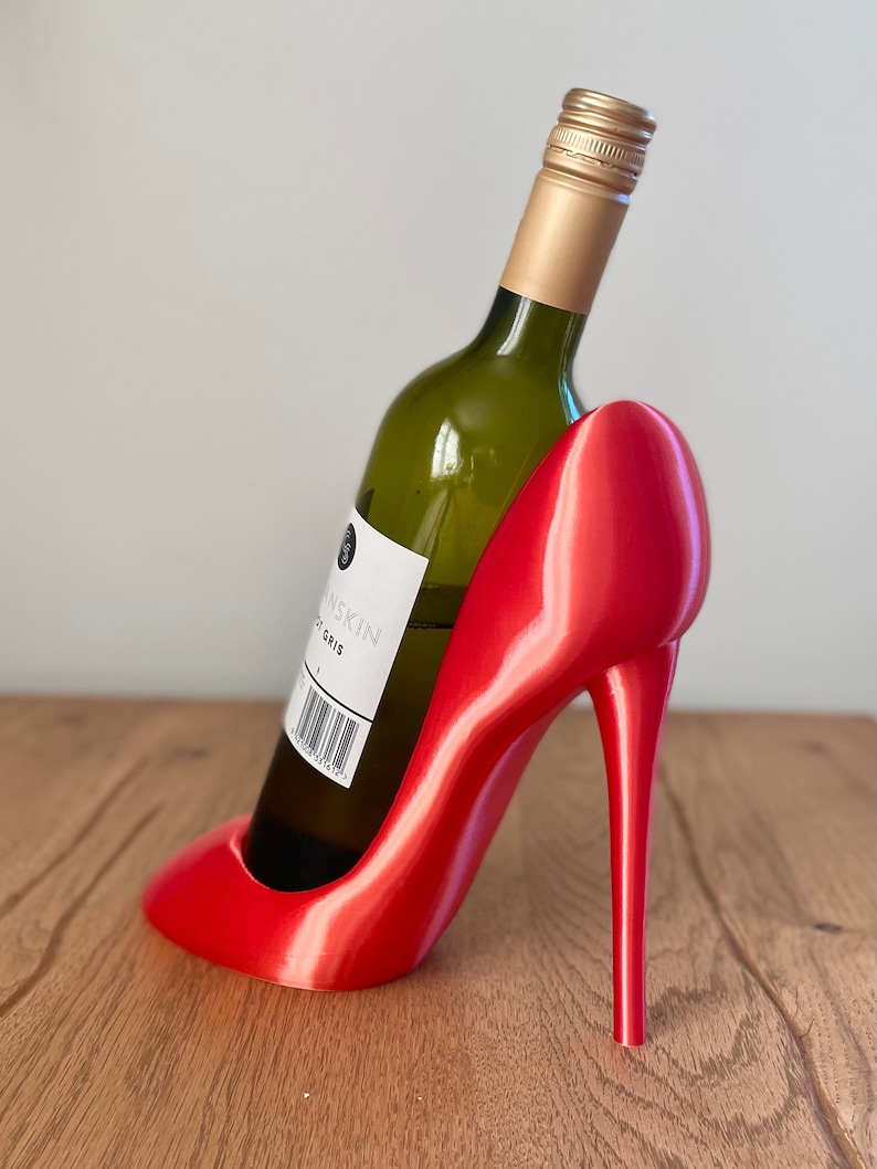 May include: A red high heel shoe shaped wine bottle holder. A bottle of white wine is in the holder.