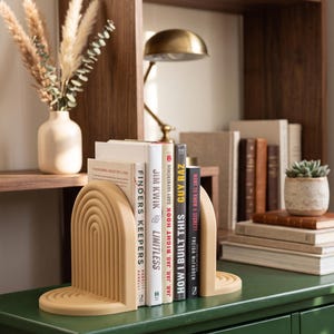 Op de afbeelding: Beige boogvormige boekensteunen met een gelaagd ontwerp, die boeken op een groen oppervlak vasthouden. Titels zijn onder meer "Finders Keepers" en "How I Built This". Een kleine vetplant en een lamp zijn zichtbaar op de achtergrond.