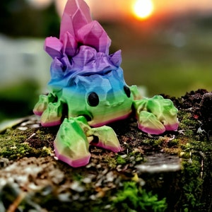 Op de afbeelding: Een regenboogkleurig, 3D-geprint beeldje van een wezen met een kristalvormig hoofd en meerdere poten. Het beeldje staat op een mossig oppervlak.