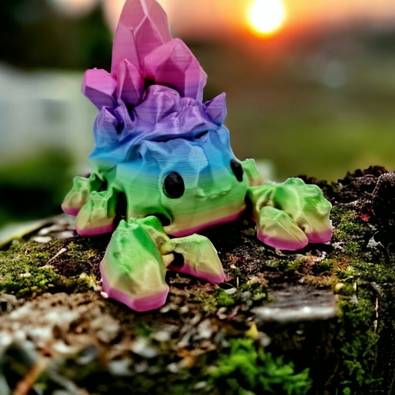 Op de afbeelding: Een regenboogkleurig, 3D-geprint beeldje van een wezen met een kristalvormig hoofd en meerdere poten. Het beeldje staat op een mossig oppervlak.