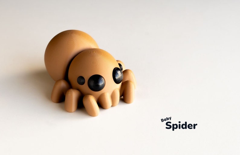 May include: A brown, 3D printed, cartoon spider with large black eyes. The spider is sitting on a white surface. The text "Baby Spider" is written below the spider.