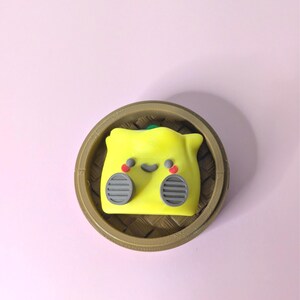 May include: A yellow dumpling character with a smiling face, red accents, and gray details, sits in a brown bamboo steamer. The dumpling has a green top and gray side details. The background is a solid light pink.