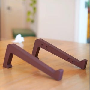 Peut inclure: Deux supports pour ordinateur portable bruns sont inclin&eacute;s sur une surface en bois. Chaque support a une base rectangulaire et un support inclin&eacute; avec trois petits trous. Les supports sont con&ccedil;us pour sur&eacute;lever un ordinateur portable.