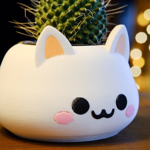 May include: White ceramic planter shaped like a cat's face with pink ears, black eyes, and a pink nose. The planter has a small cactus plant inside.
