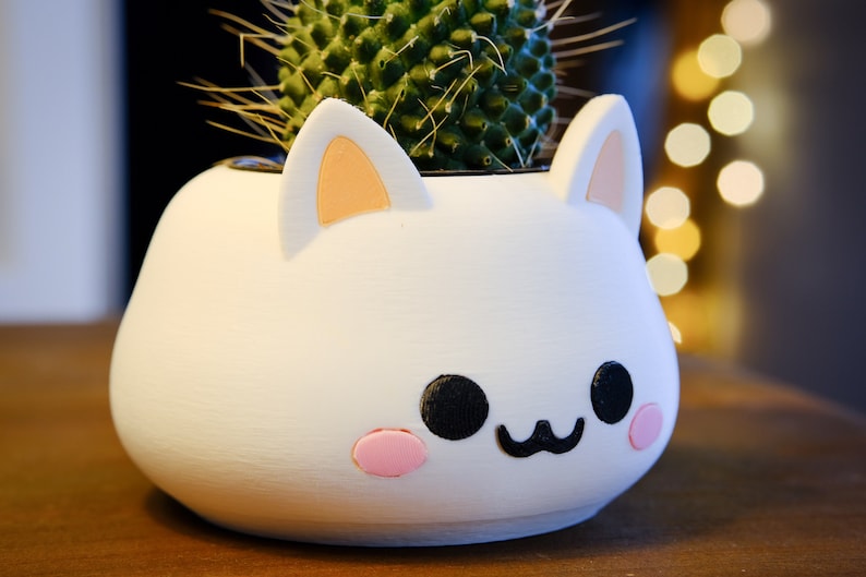May include: White ceramic planter shaped like a cat's face with pink ears, black eyes, and a pink nose. The planter has a small cactus plant inside.