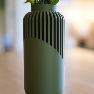 May include: A green vase with a unique design, featuring vertical grooves on the upper portion and a curved, smooth section on the lower part. The vase holds white flowers, creating a contrast with the matte green finish. A modern home decor item.