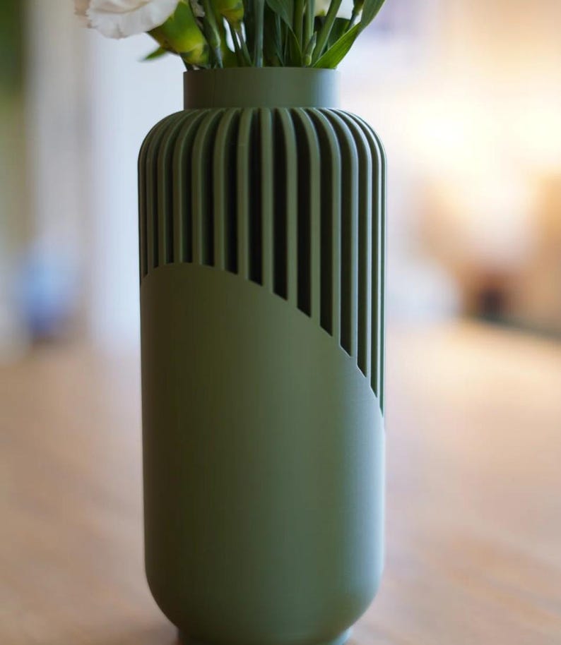 May include: A green vase with a unique design, featuring vertical grooves on the upper portion and a curved, smooth section on the lower part. The vase holds white flowers, creating a contrast with the matte green finish. A modern home decor item.