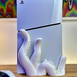 May include: A white PlayStation 5 console with a 3D printed white octopus stand. The octopus tentacles wrap around the console and have suction cups.