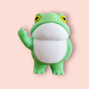 May include: A green frog figurine with yellow eyes and a white belly. The frog is giving the middle finger.
