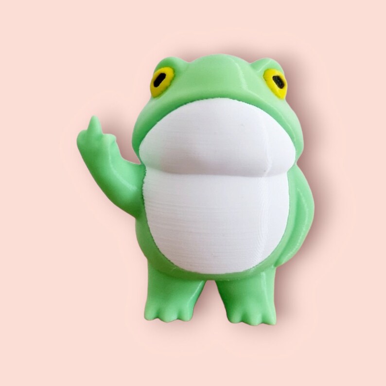 May include: A green frog figurine with yellow eyes and a white belly. The frog is giving the middle finger.