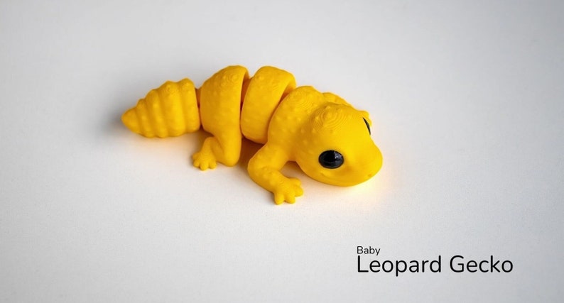 May include: A yellow plastic toy of a baby leopard gecko with segmented body parts. The toy has black eyes and a textured surface.