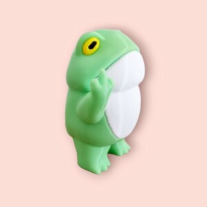 May include: A green frog figurine with a white belly and a yellow eye. The frog is standing on its hind legs with its front legs raised.