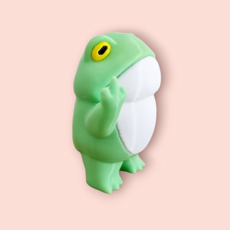 May include: A green frog figurine with a white belly and a yellow eye. The frog is standing on its hind legs with its front legs raised.