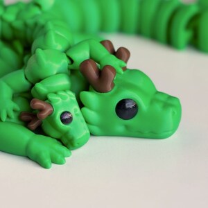 May include: A green dragon figurine with brown antlers, holding a smaller green dragon figurine with brown antlers. The dragons have large, black eyes and are lying down.