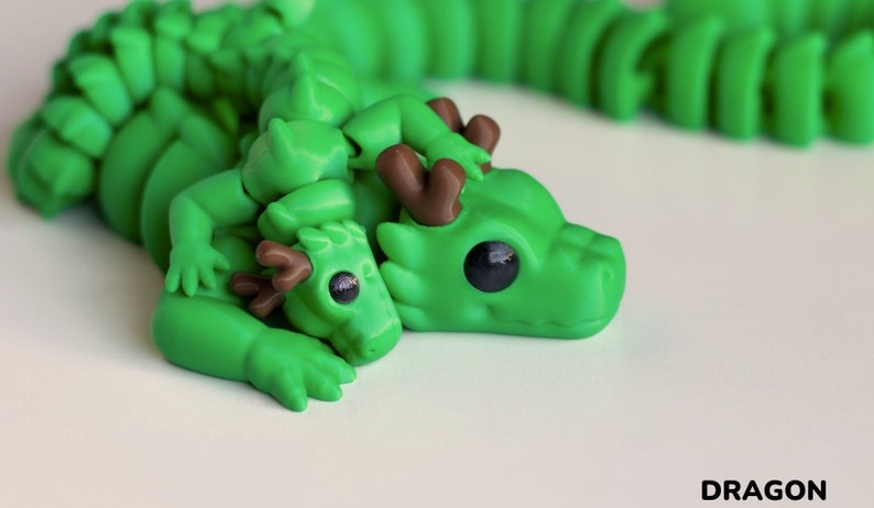 May include: A green dragon figurine with brown antlers, holding a smaller green dragon figurine with brown antlers. The dragons have large, black eyes and are lying down.