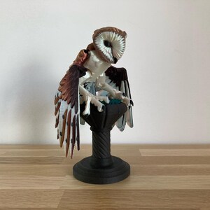 May include: A white and brown owl figurine with a black base. The owl is perched on a pedestal with a round top and a fluted base. The owl's wings are spread out and its eyes are closed.