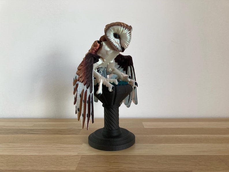 May include: A white and brown owl figurine with a black base. The owl is perched on a pedestal with a round top and a fluted base. The owl's wings are spread out and its eyes are closed.