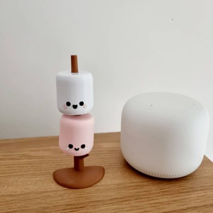 Op de afbeelding: Een witte en roze marshmallow-figuur met een glimlachend gezicht, gestapeld op een bruine stok, op een houten oppervlak. Een witte slimme speaker staat op de achtergrond.