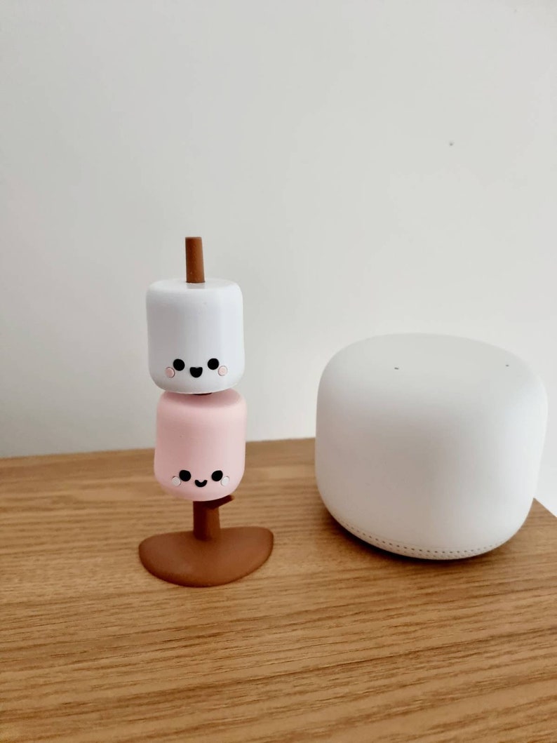 Op de afbeelding: Een witte en roze marshmallow-figuur met een glimlachend gezicht, gestapeld op een bruine stok, op een houten oppervlak. Een witte slimme speaker staat op de achtergrond.