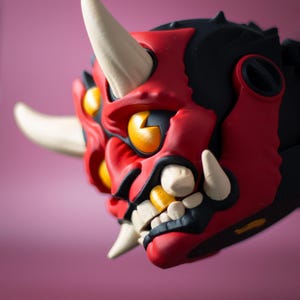 May include: A detailed, close-up view of a red, black, and yellow Oni mask. The mask features white horns and teeth, with yellow eyes and a fierce expression. The mask is a striking piece of art.