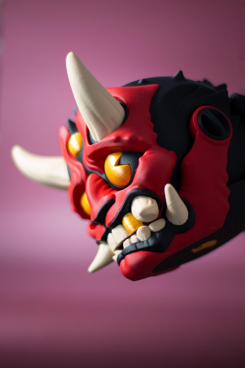 May include: A detailed, close-up view of a red, black, and yellow Oni mask. The mask features white horns and teeth, with yellow eyes and a fierce expression. The mask is a striking piece of art.